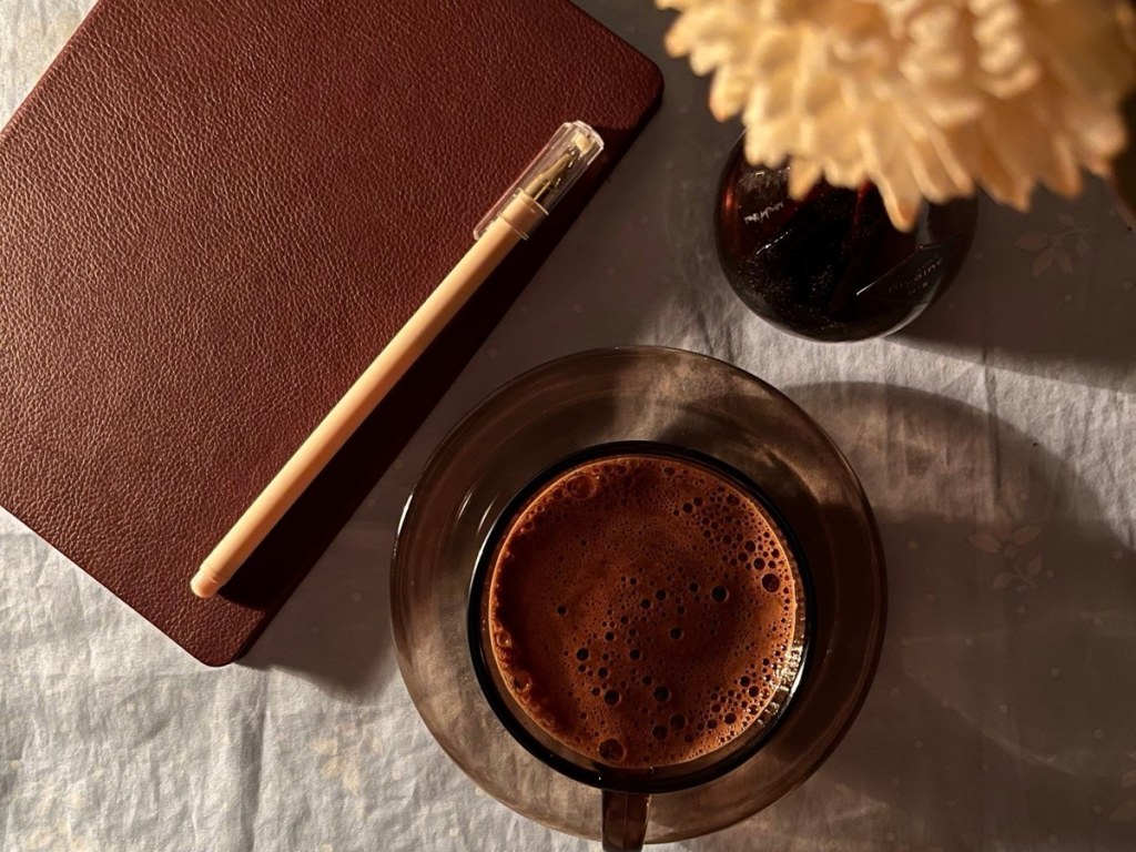 Brown notebook with a pen next to a cup of coffee and cream flowers on a linen tablecloth